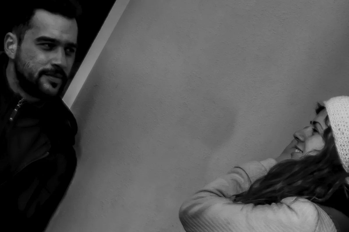Dark-haired man peering around a wall toward a smiling woman in a knit hat